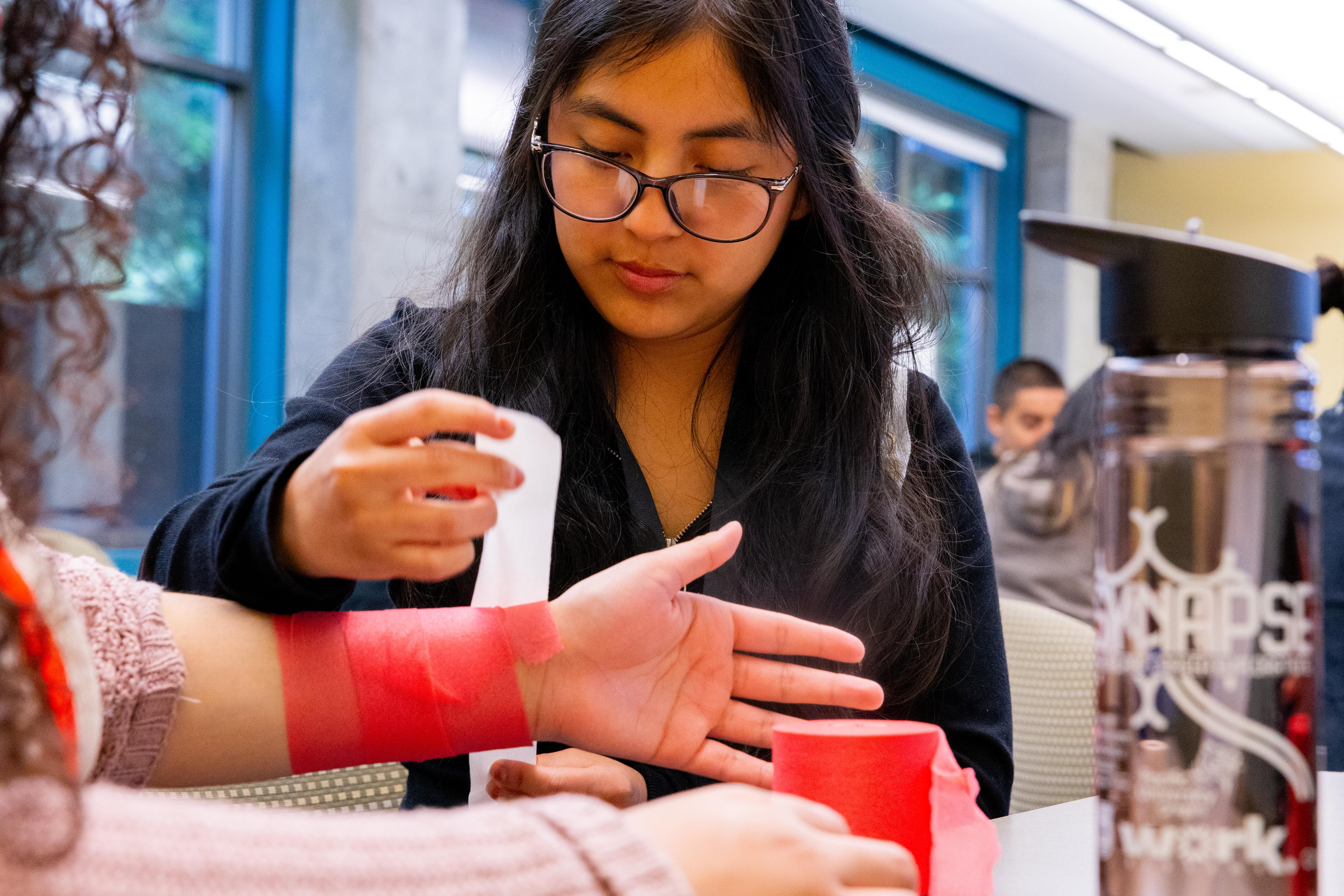 A student trains by placing a bandage on another student's wrist.