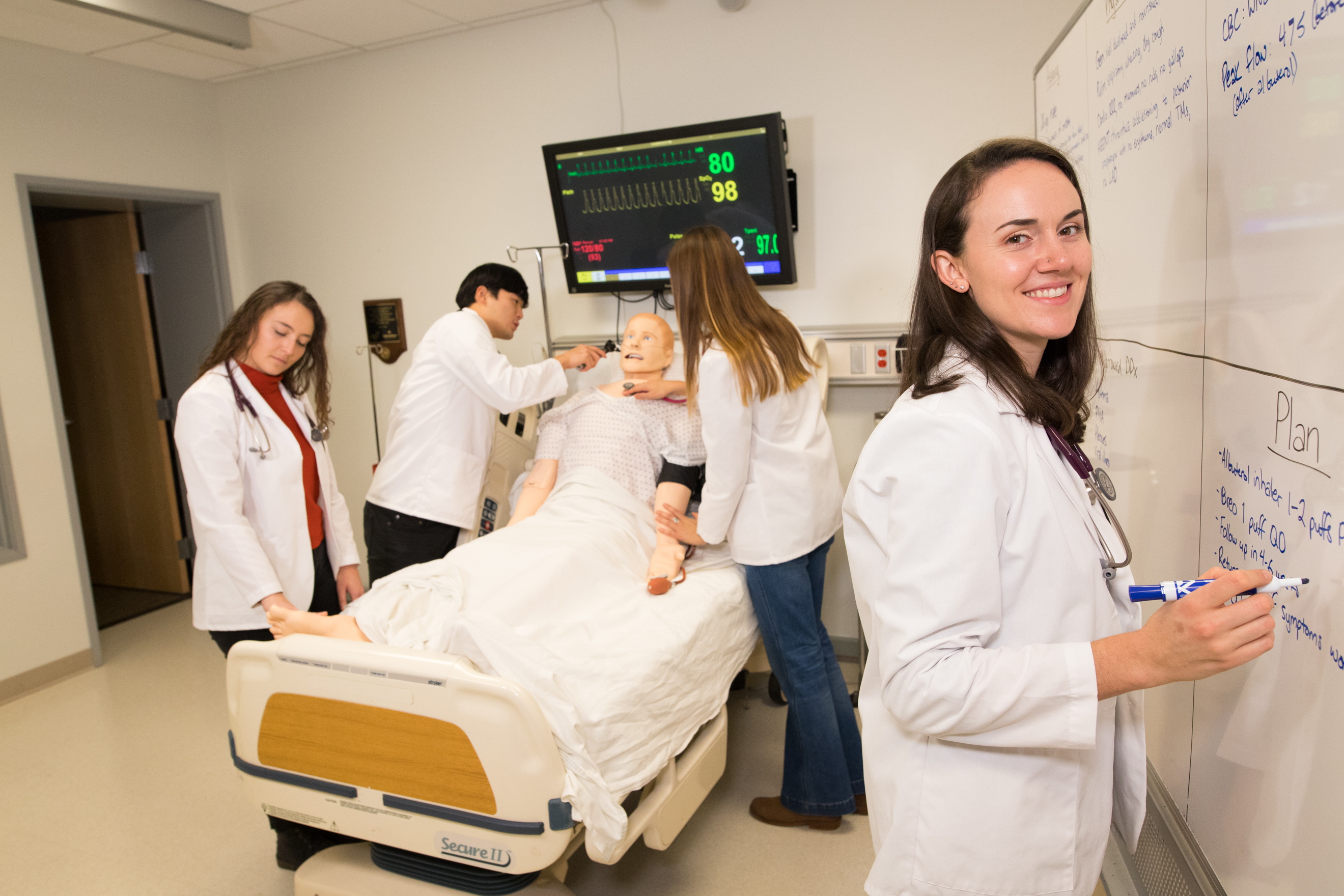 Four physician assistant program students demonstrate care on a dummy body lying in a hospital bed.
