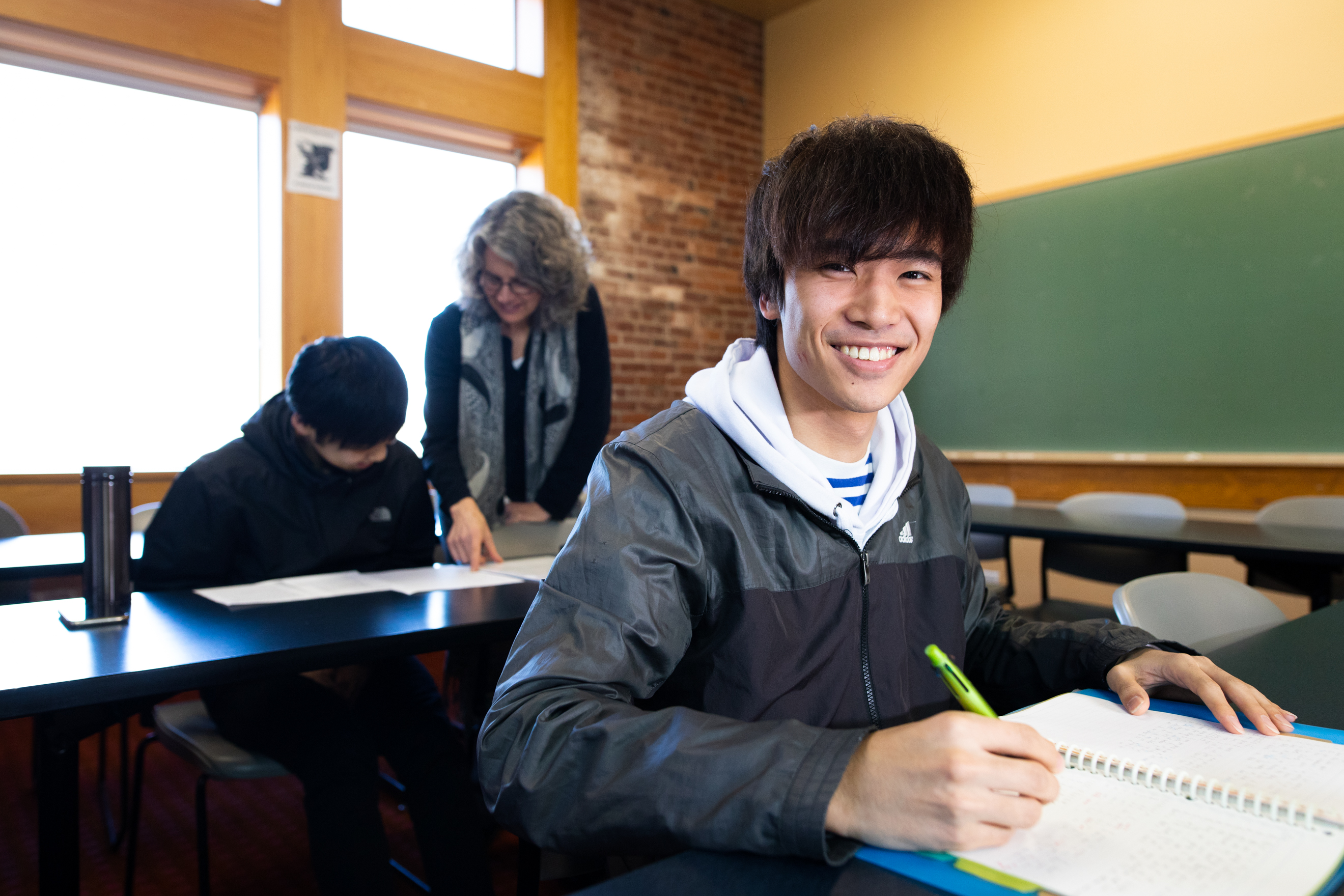 A student in a classroom looks into the camera between taking notes during a class.