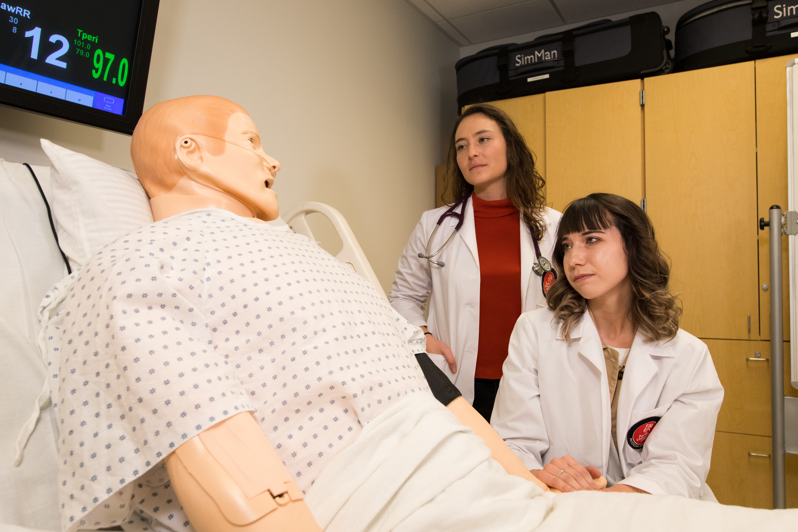 Two female physician assistant studies students observe a training dummy.