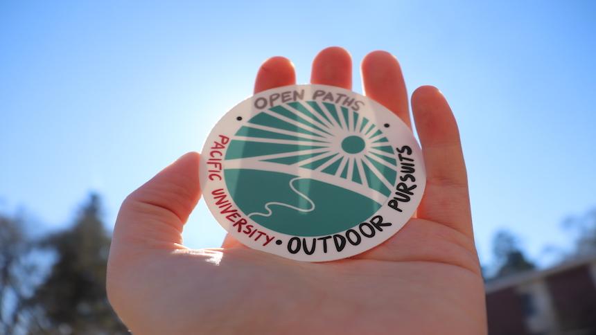 A student's hand holds an Outdoor Pursuits Open Paths sticker up to the sunlight. 