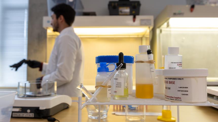 A pharmacy student walks in the background while a set of pharmacy chemicals sits in the foreground. 
