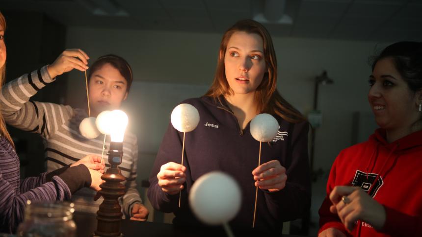 A female school teacher facilitates a demonstration model of the solar system for their students.