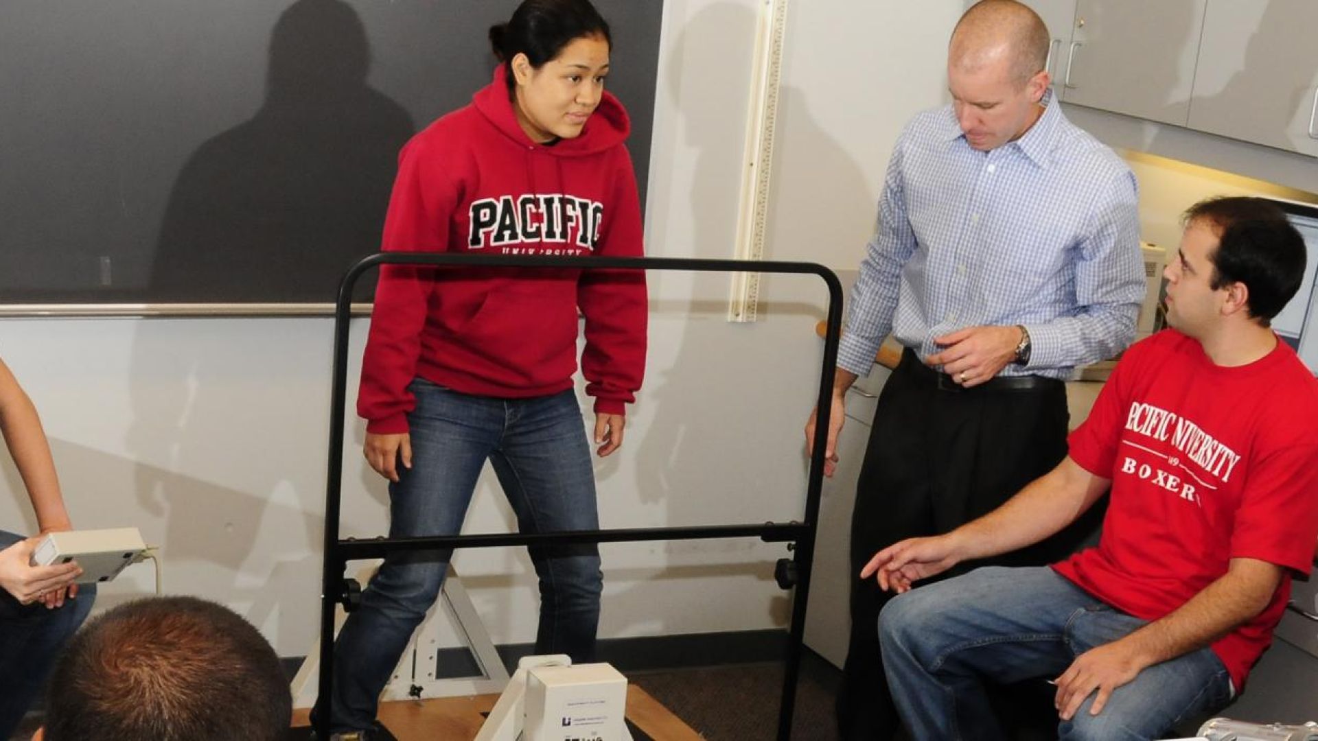 Students sitting and standing in room, one standing on piece of equipment