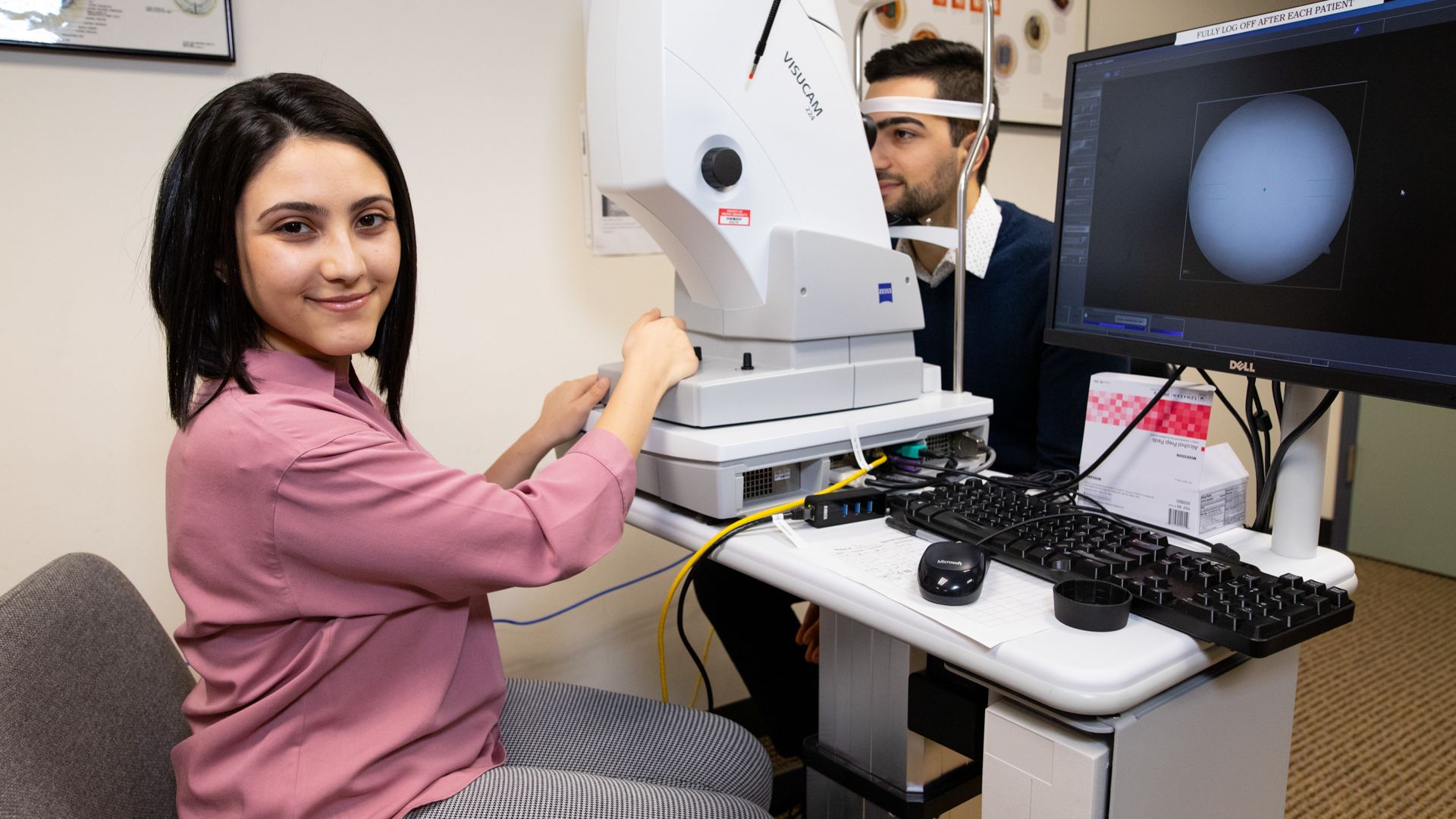 Woman using a retinal camera on someone, turned to camera and smiling